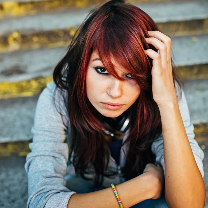Young woman in a hoodie with long red hair sitting outside on steps, stares into the camera looking serious; she holds her long red hair over part of her face to hide bruises