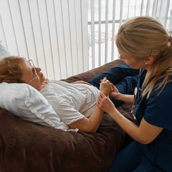 Female in teal scrubs holds the hand of an older female patient while sitting at her bedside