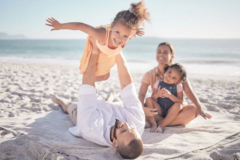 Attractive young family sit on a blanket on the beach while dad playfully holds his daughter in the air