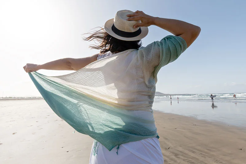 Back view of brunette with thin scarf and straw hat enjoying freedom while spending windy day on sandy beach