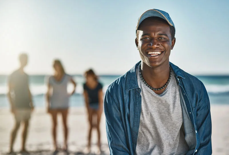 Young adult African American man on the beach in a ball cap smiles broadly at the camera while friends stand in the background