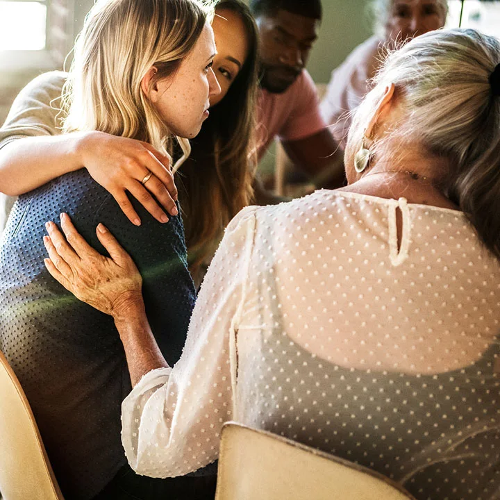 people sitting in a circle of chairs show support to young blonde woman, touching her shoulder