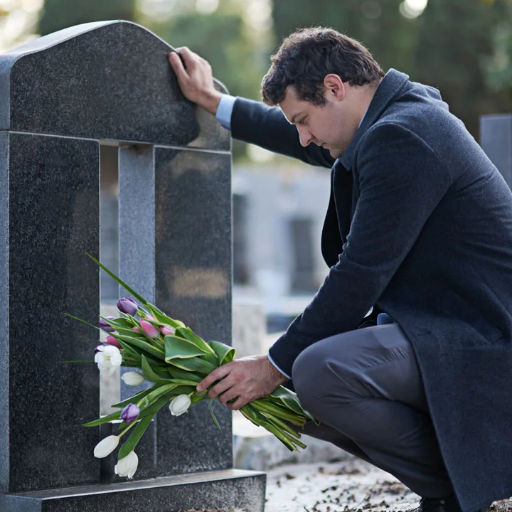 man holding flowers stands at a gravesite with his hand on the tombstone, as if in mourning