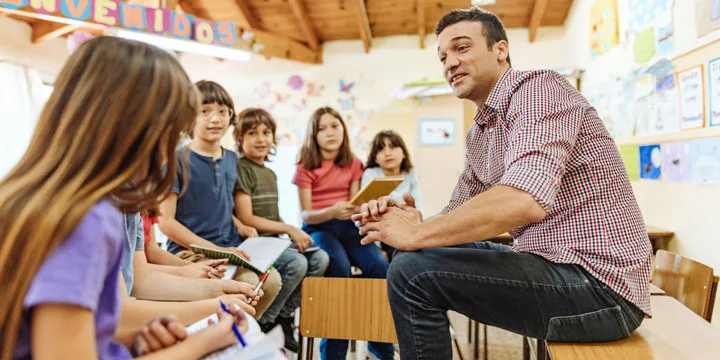 male teacher dressed casually sits, speaking to young students