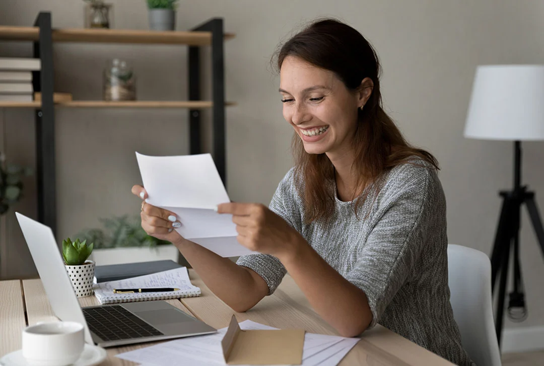Woman sitting at a desk with her laptop opens a letter and has a huge, excited smile