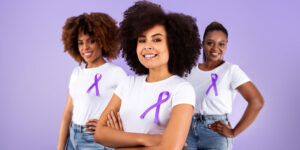 Three women stand confidently against a light purple background, each wearing a white T-shirt with a purple awareness ribbon pinned to the front, symbolizing support for domestic violence awareness.