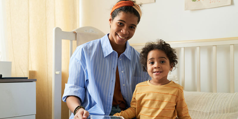Young black woman in a blue shirt sits on a daybed with a young child, both smiling