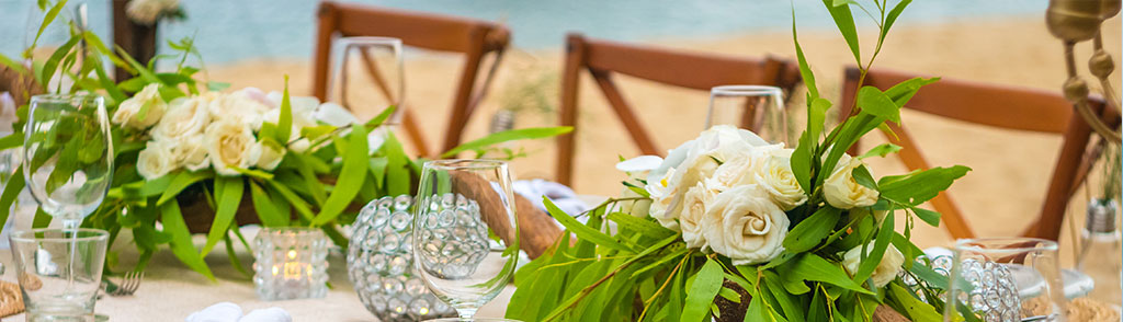Elegant outdoor table setting with white rose centerpieces, greenery, glassware, and candles arranged near a sandy beach with water in the background