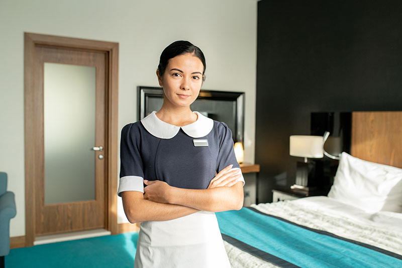 A young Latina housekeeper in uniform stands in front of a hotel bed with arms crossed