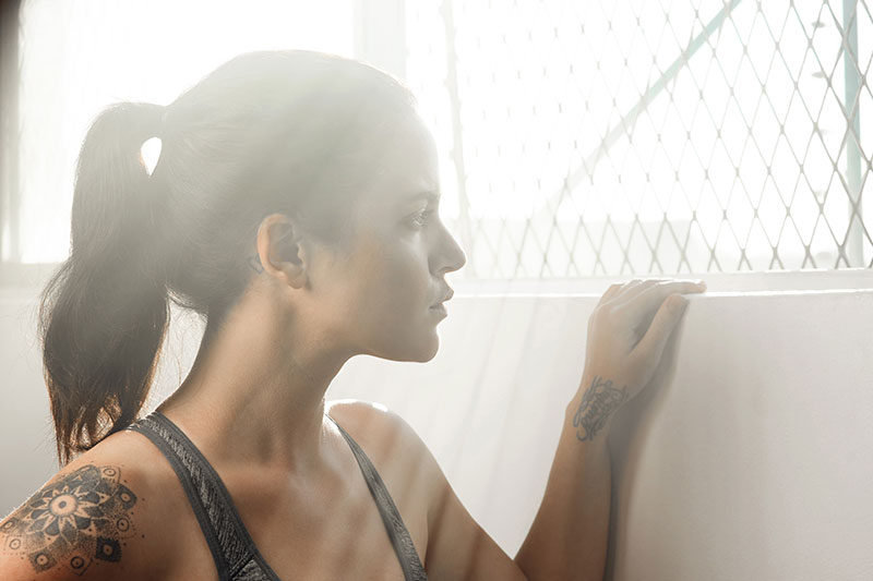 A young woman with tattoos looks pensive as she stares out through a chain fence