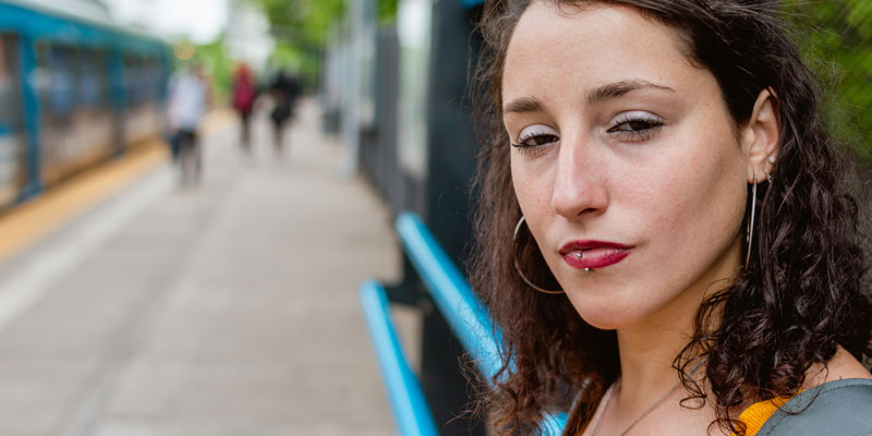 A femme person with brown curly hair and piercings stands outside while others walk by, looking seriously at the camera