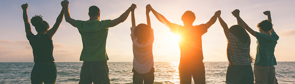 Silhouettes of six people standing side by side on a beach at sunset, holding hands raised high in a gesture of unity and celebration.