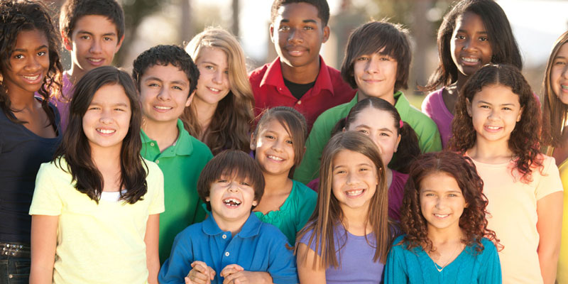 A group of fifteen children of diverse ages and races, all wearing brightly colored shirts of different colors, smiling