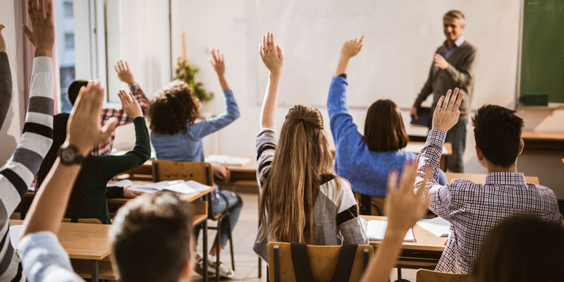 A male teacher at the head of a classroom of young teens, with several students raising their hands