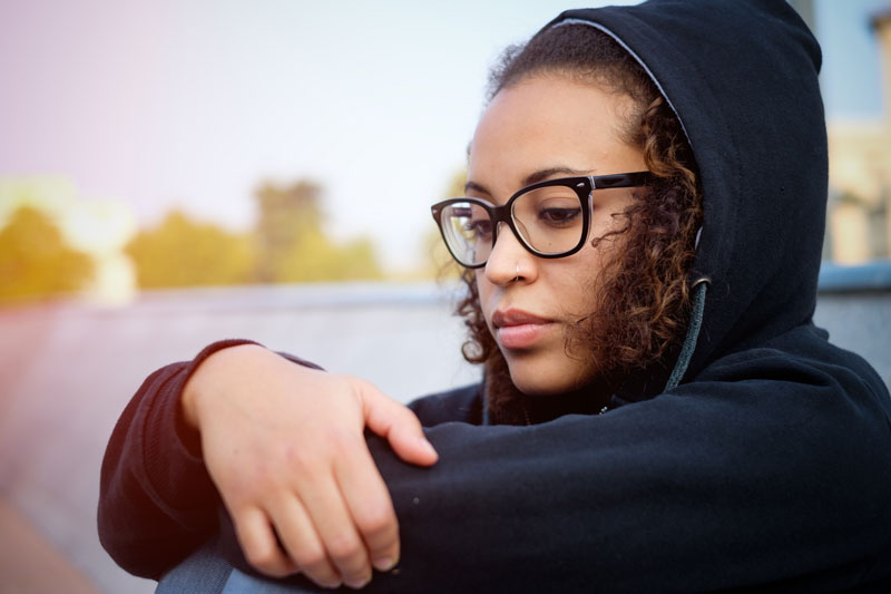 Young woman with curly brown hair in glasses and a navy hoodie sits with her knees pulled up and her arms folded, looking as if she's thinking seriously