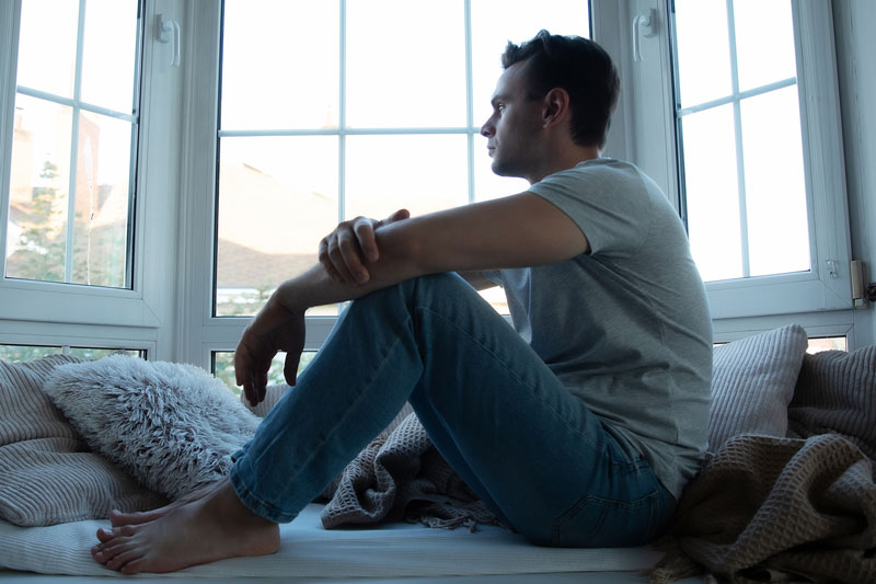 Young male sits on a bed in front of a row of windows, looking out the windows as if lost in his thoughts