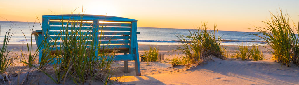 A weathered blue wooden bench sits on a sandy beach with setting sun over the ocean in the background