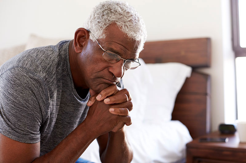 Older black man with gray hair sits on the edge of his bed with hands clasped as if deep in thought