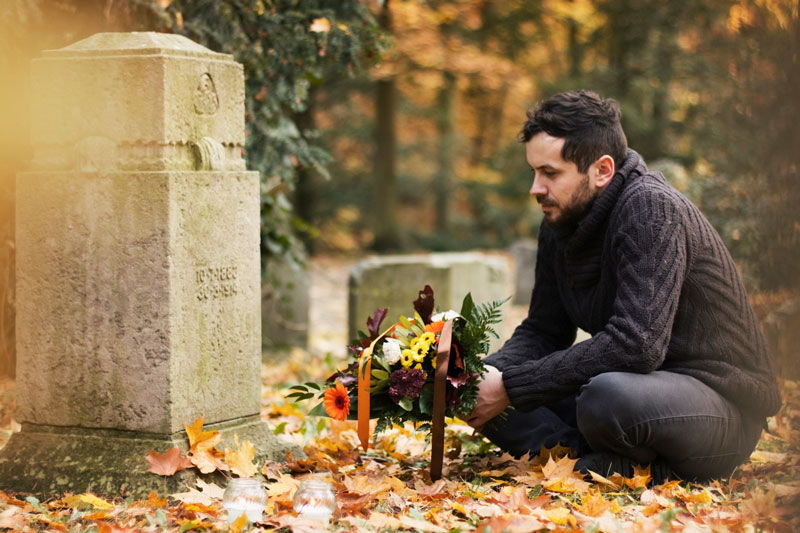 Man with dark hair sits in front of a tombstone holding a bouquet of flowers